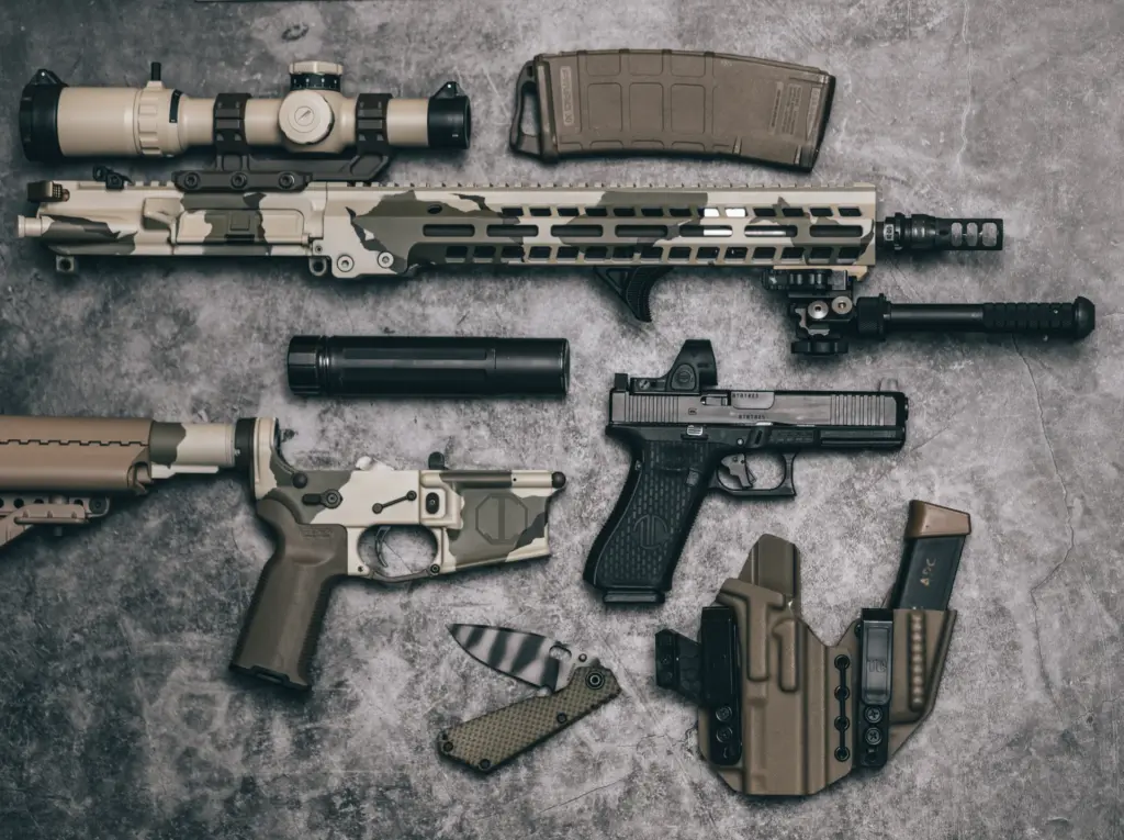 AR-15 rifle components and handgun flat-lay with optic, suppressor, magazines and holster arranged on concrete.