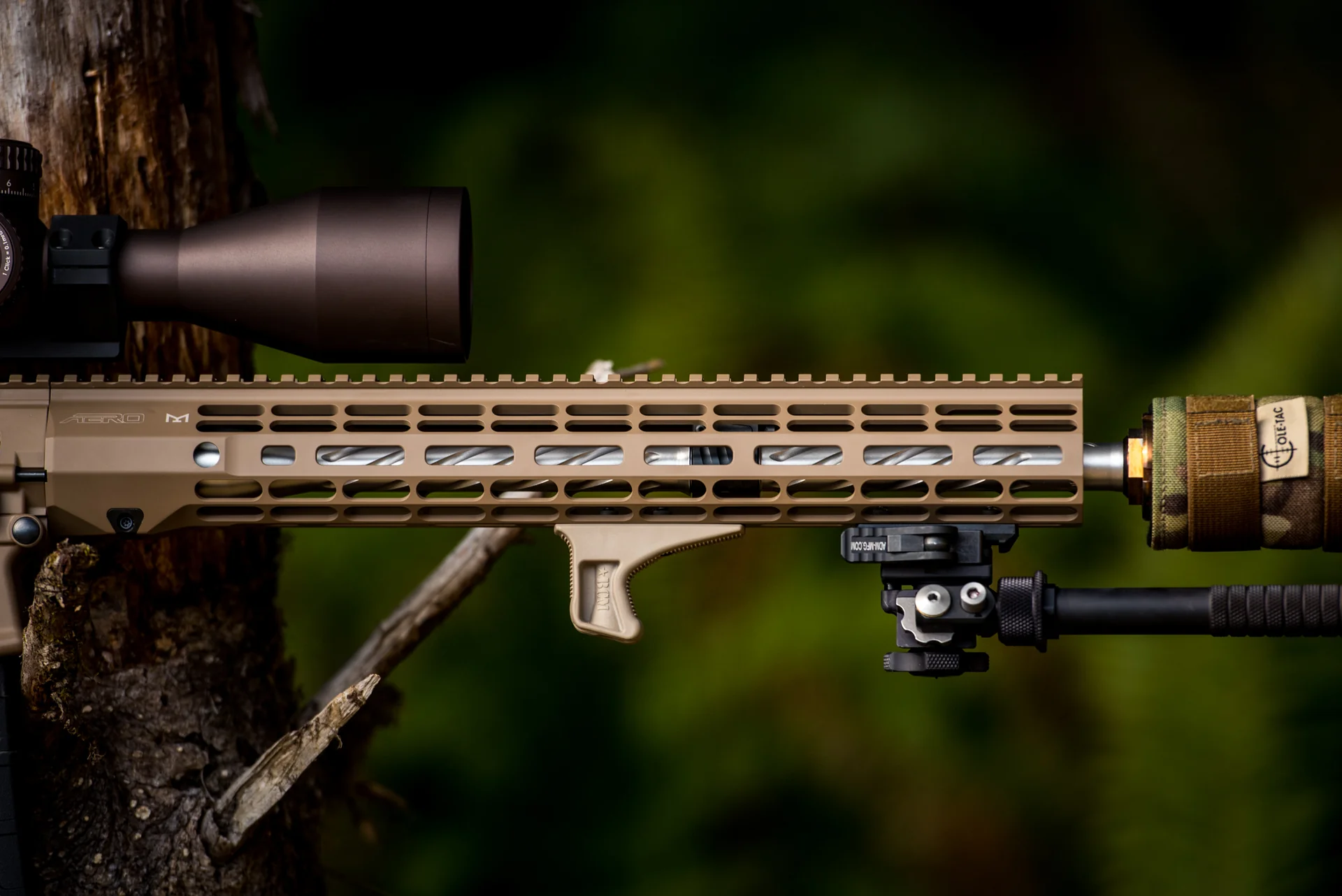 Atlas bipod deployed close-up under an FDE Aero handguard with suppressor and Ois-Tac cover against green forest.