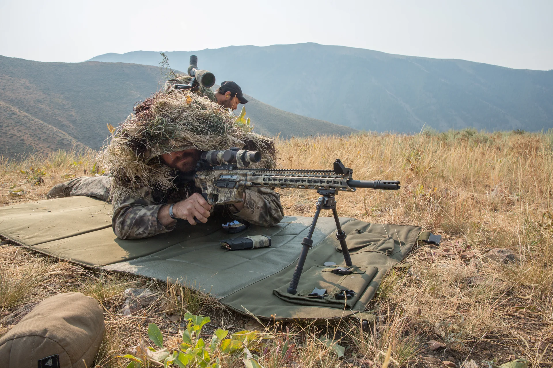 Atlas bipod deployed under an AR precision rifle as a ghillie-clad shooter takes a prone position in mountain terrain.