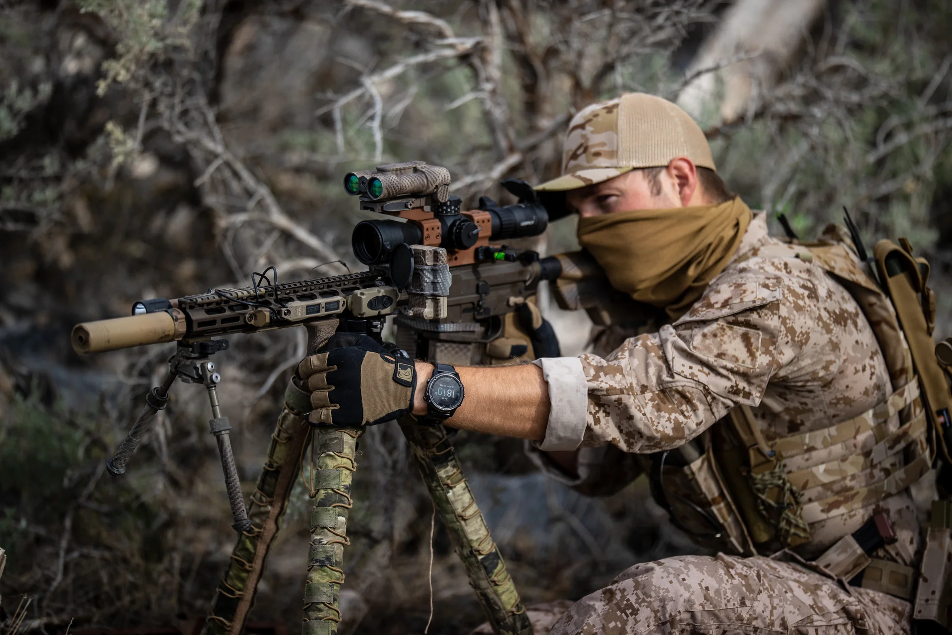 Atlas bipod deployed under a tan AR-style rifle as a kneeling operator aims through a scope in desert brush.