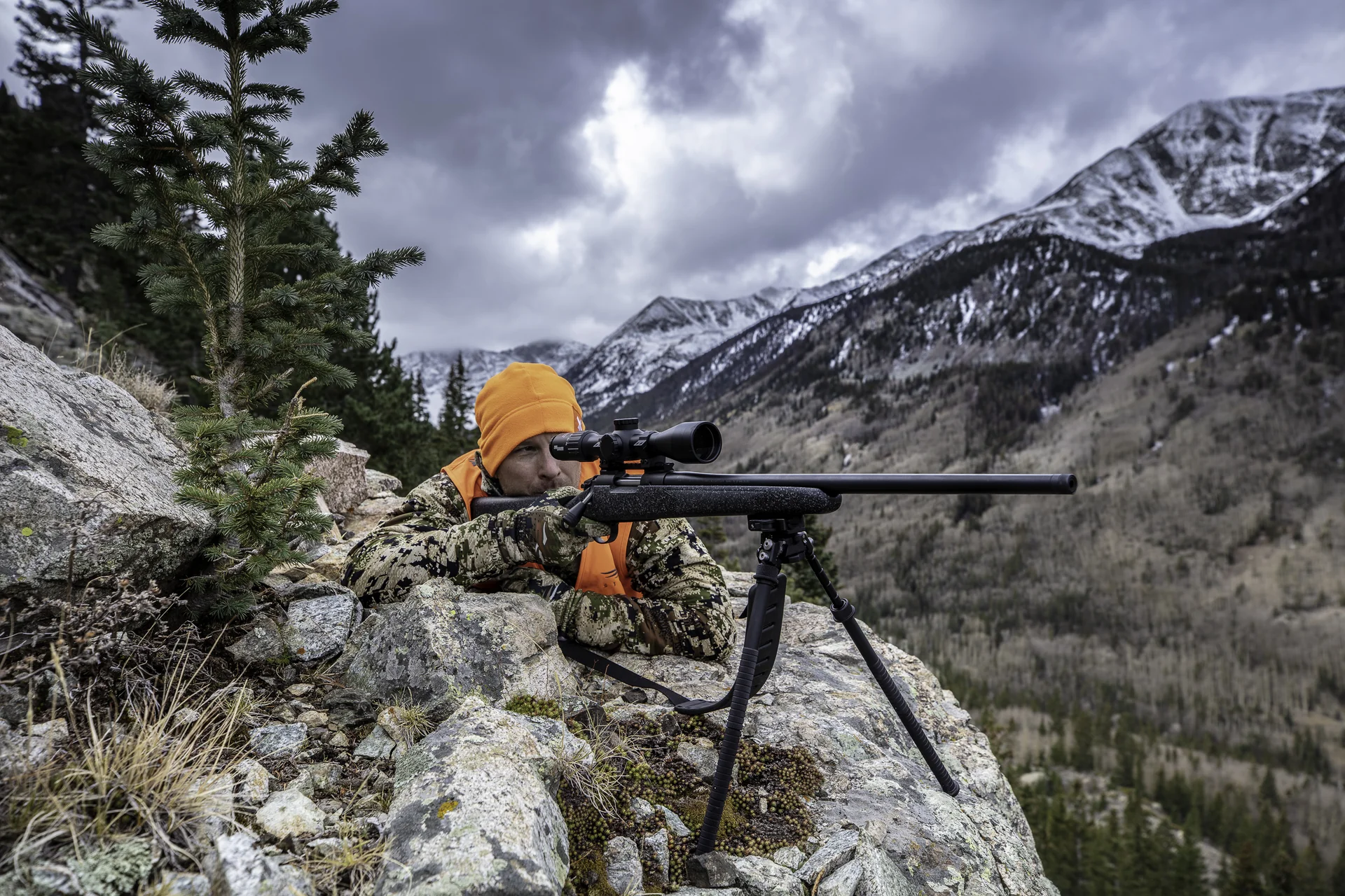 Atlas bipod deployed under a bolt-action rifle as a prone hunter aims from a rocky cliff toward mountains.