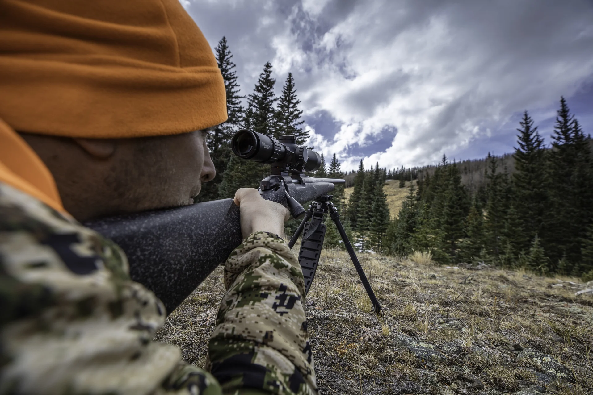 Atlas bipod deployed under a bolt-action rifle steadied by a prone shooter glassing a stormy mountain landscape.