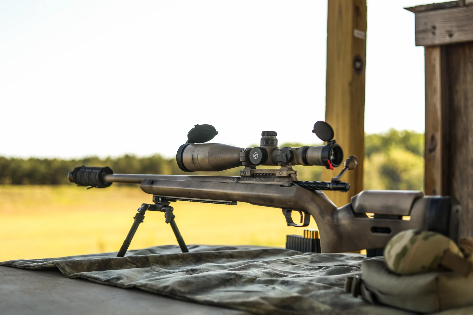 Atlas bipod deployed under a camo-finished bolt-action precision rifle resting at a range with golden field behind.