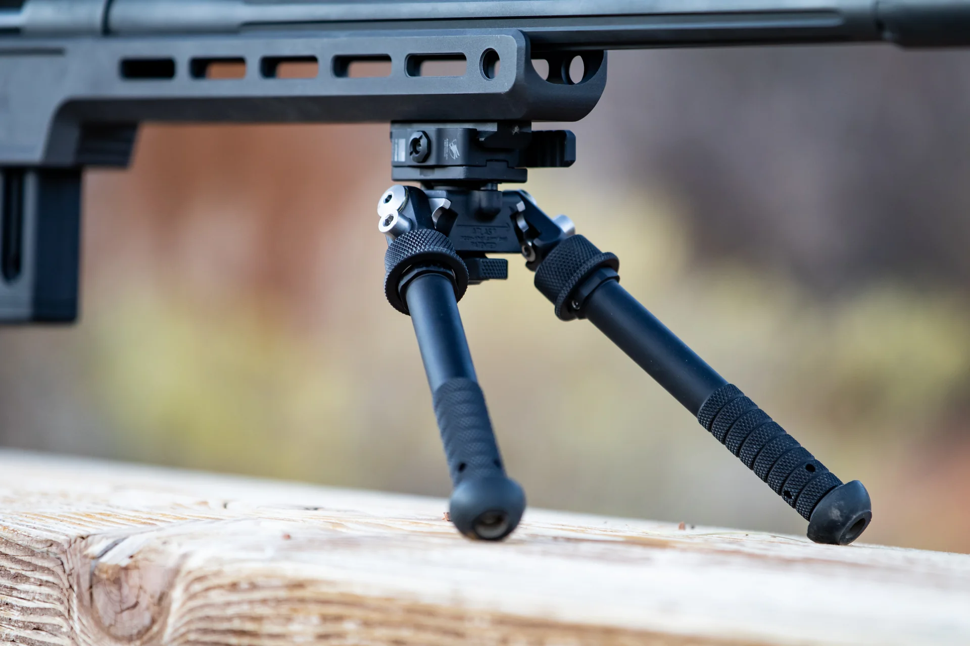 Atlas bipod deployed with splayed notched legs under a black chassis rifle resting on a wooden rail outdoors.