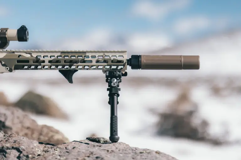 Atlas bipod deployed on the M-LOK rail of a camo suppressed rifle resting on a snowy rock outcrop.