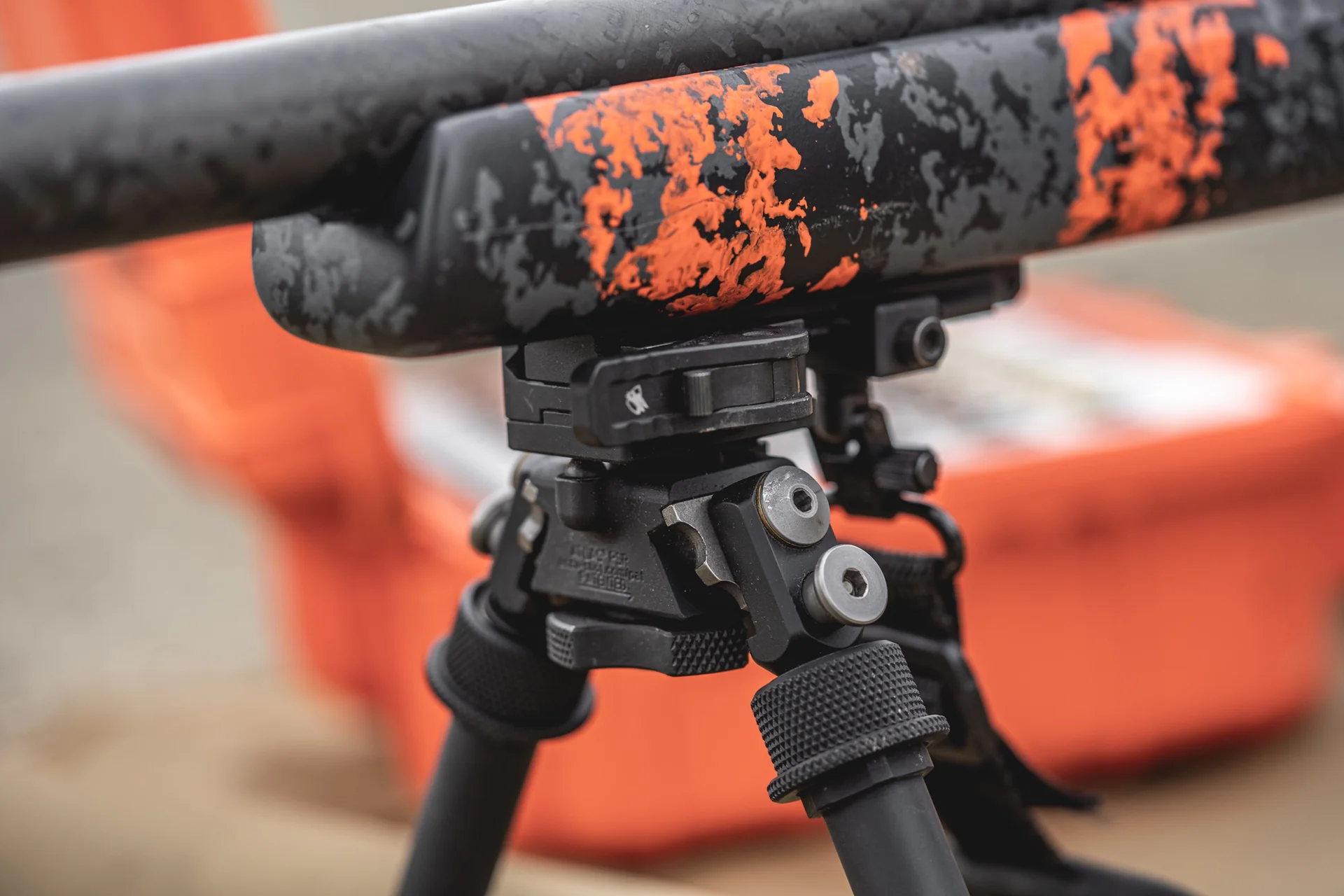 Atlas bipod deployed in close-up showing mount and notched legs under an orange-camo rifle by an orange case.