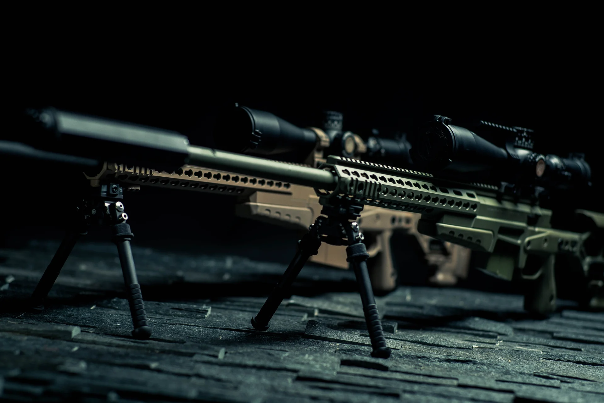 Atlas bipod deployed under a tan rail-mounted rifle beside a second FDE rifle on a dark studio table.