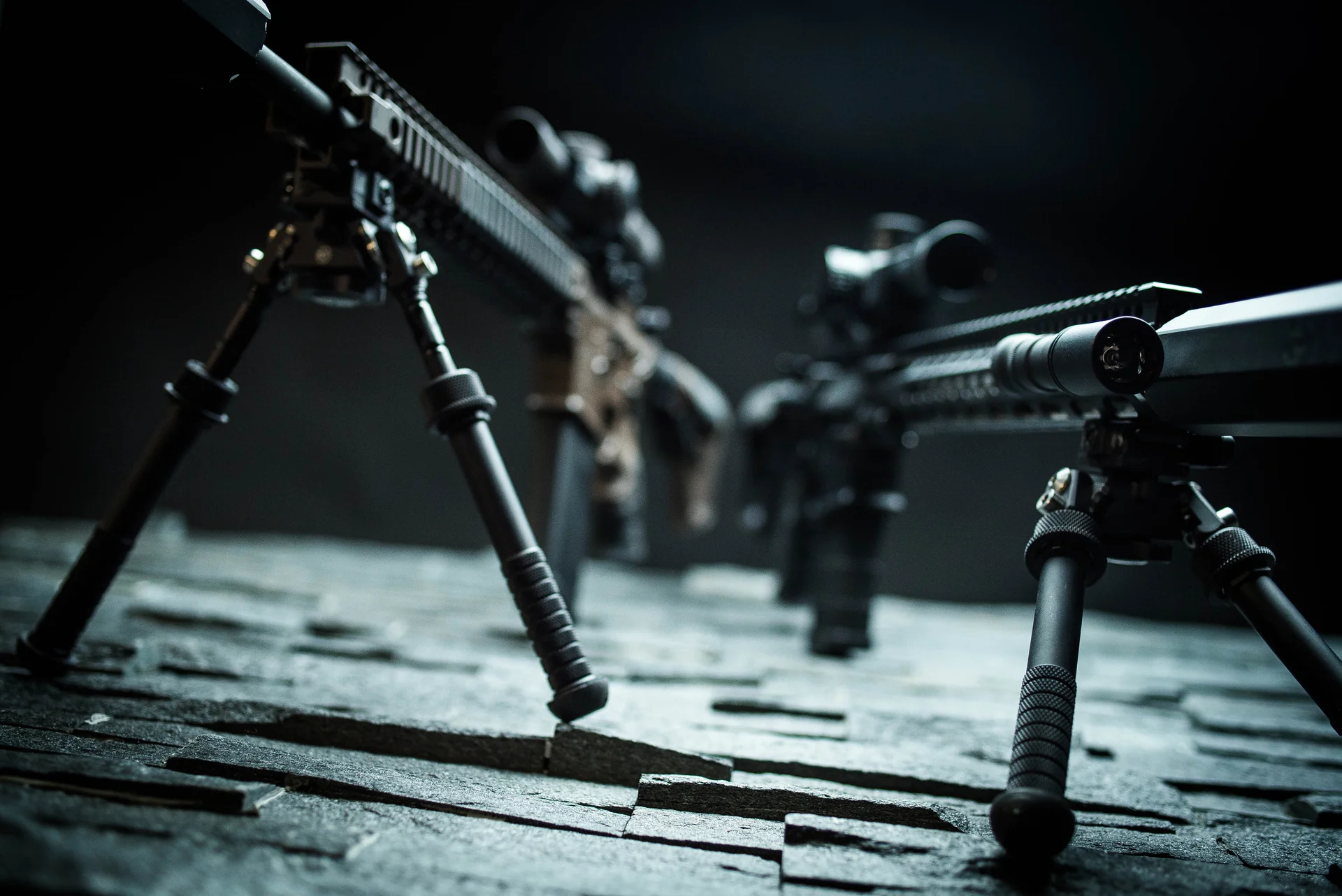 Atlas bipods deployed with splayed notched legs under two suppressed rifles in dramatic low light.