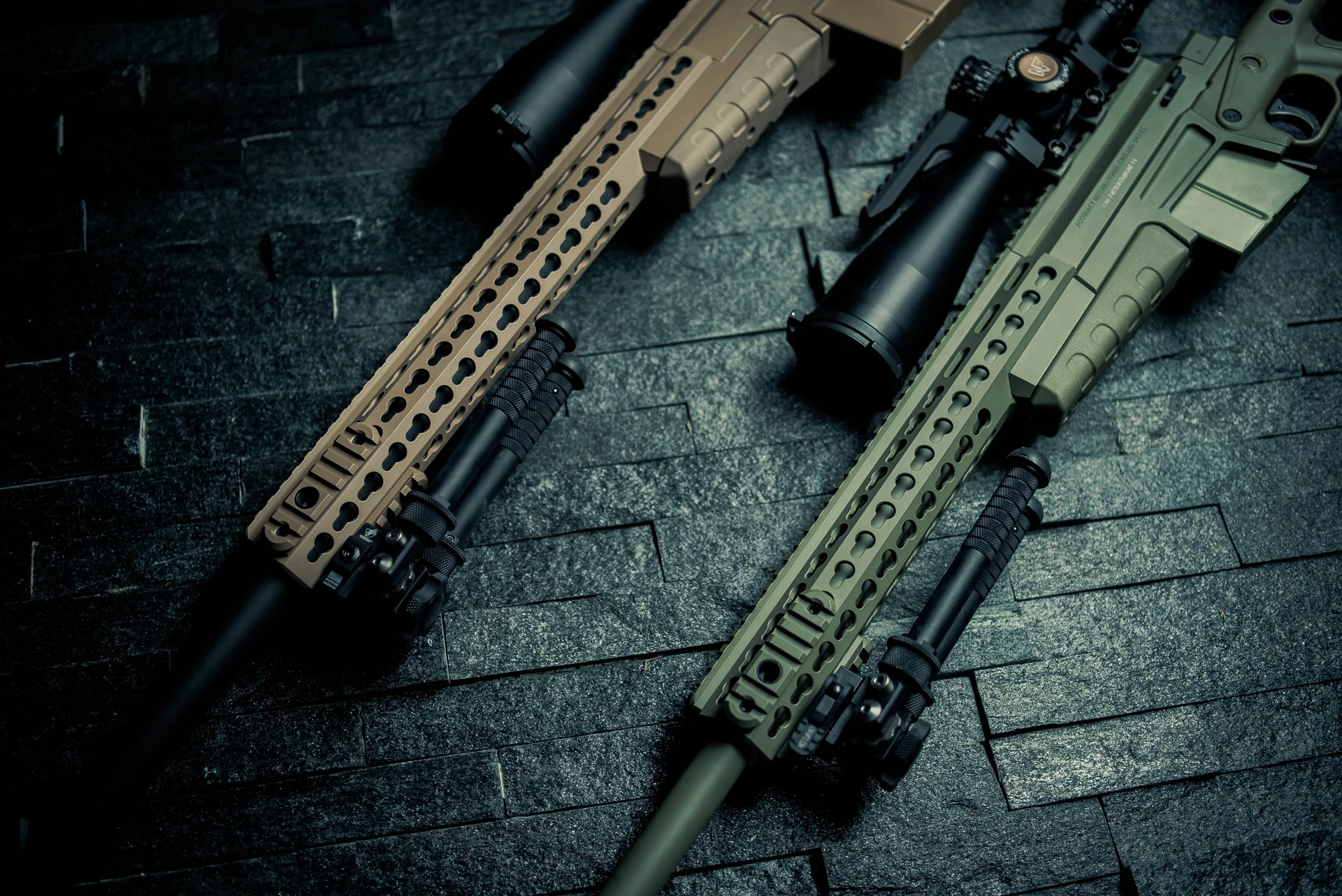 Atlas bipods deployed under two rail-mounted rifles in tan and green showing notched legs on dark slate.