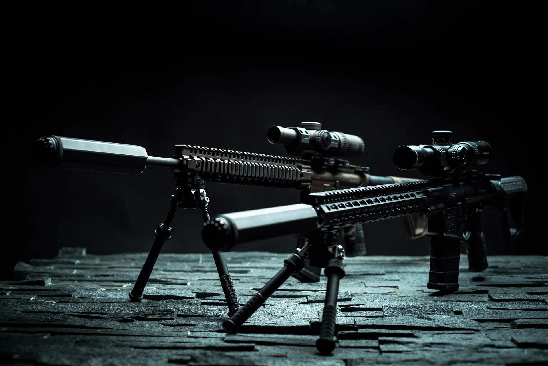 Atlas bipods deployed under two suppressed rail-mounted rifles with scopes on a dark studio table.