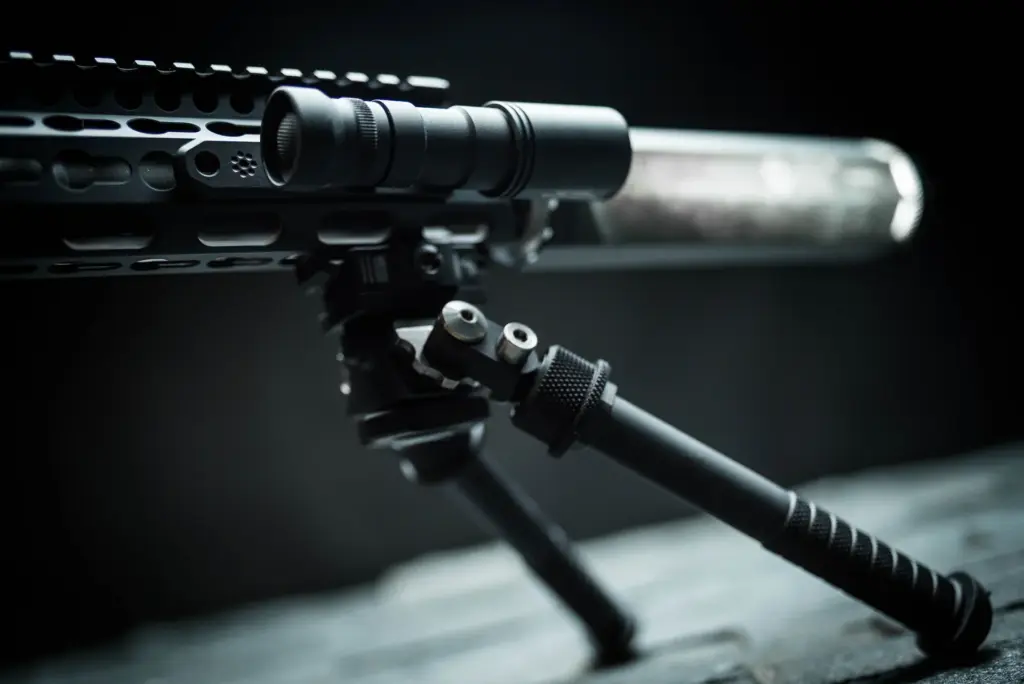 Atlas bipod deployed supporting a rail-mounted rifle equipped with a weapon-mounted flashlight on dark wood.