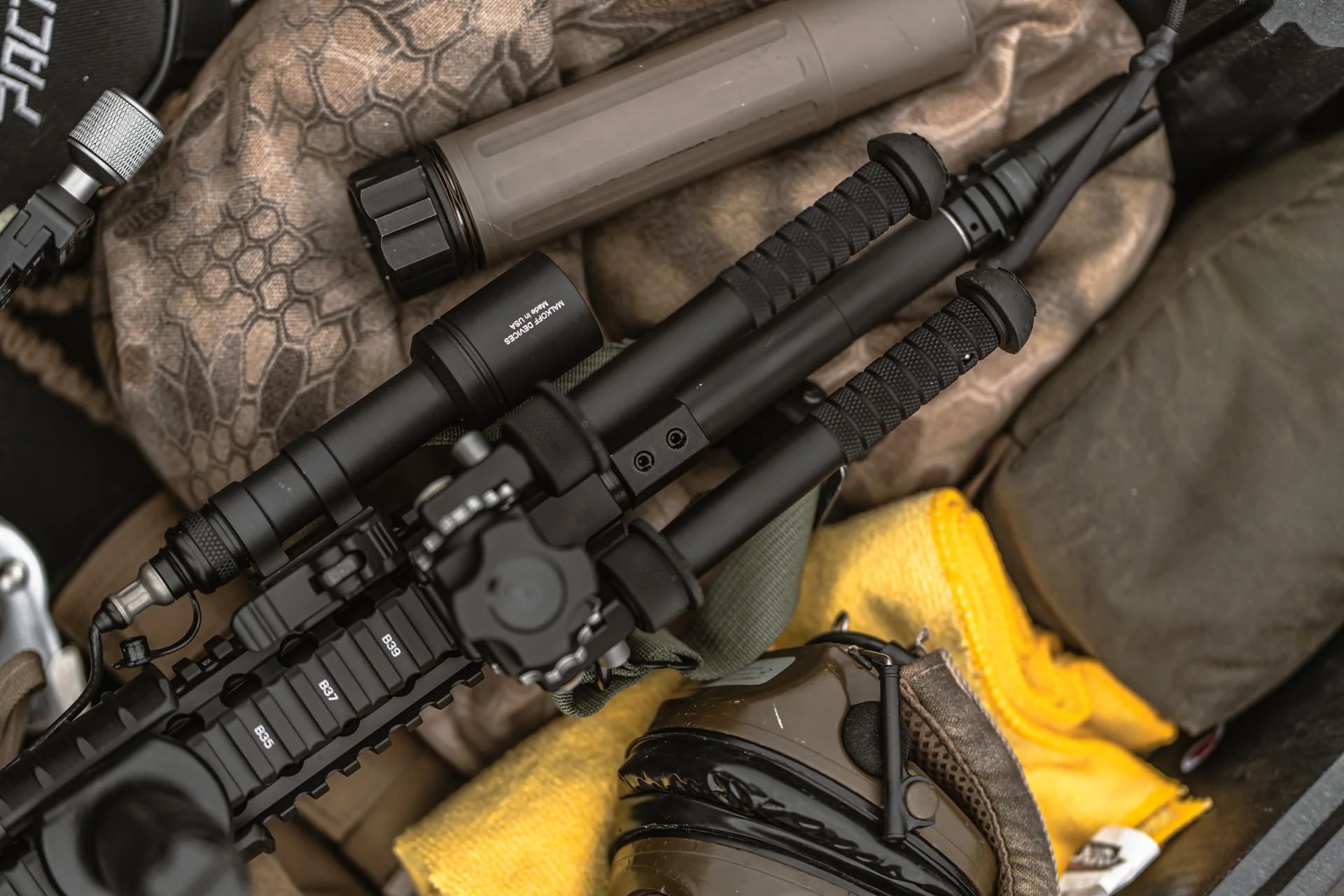 Atlas bipod folded with notched legs resting beside a suppressor on an AR-15 amid camo gear in an open range bag.