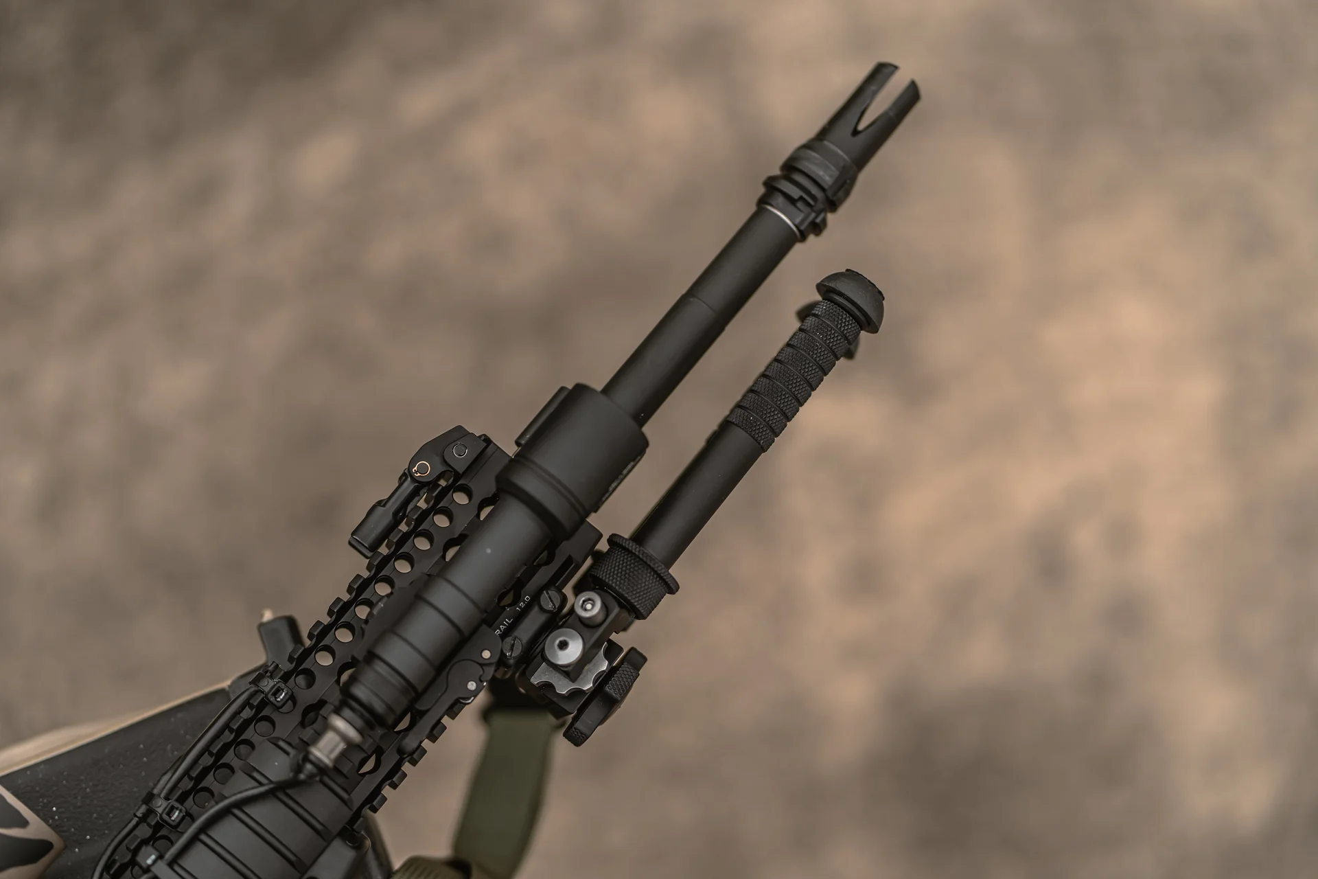 Atlas bipod folded upward with knurled notched leg against the handguard rail of an AR-15 over a sandy backdrop.