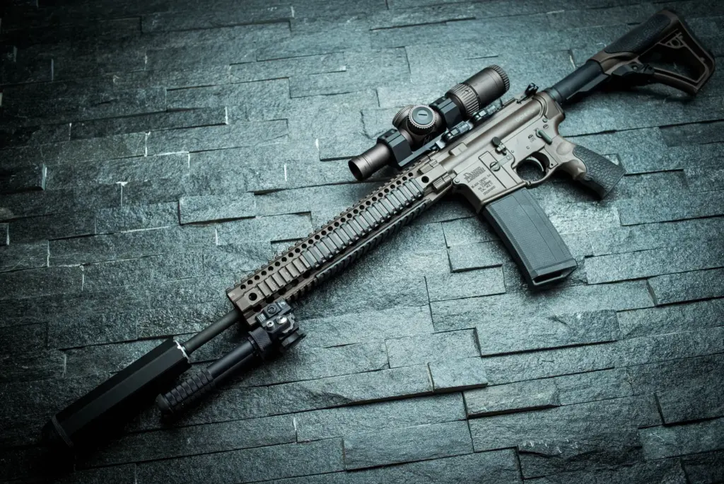 Atlas bipod folded under the fore-end of an FDE suppressed AR-style carbine laid flat on dark slate.