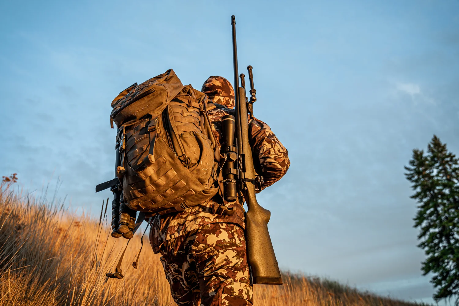 Atlas bipod folded on a shouldered hunting rifle as a camo hunter hikes a grassy hillside at dawn.
