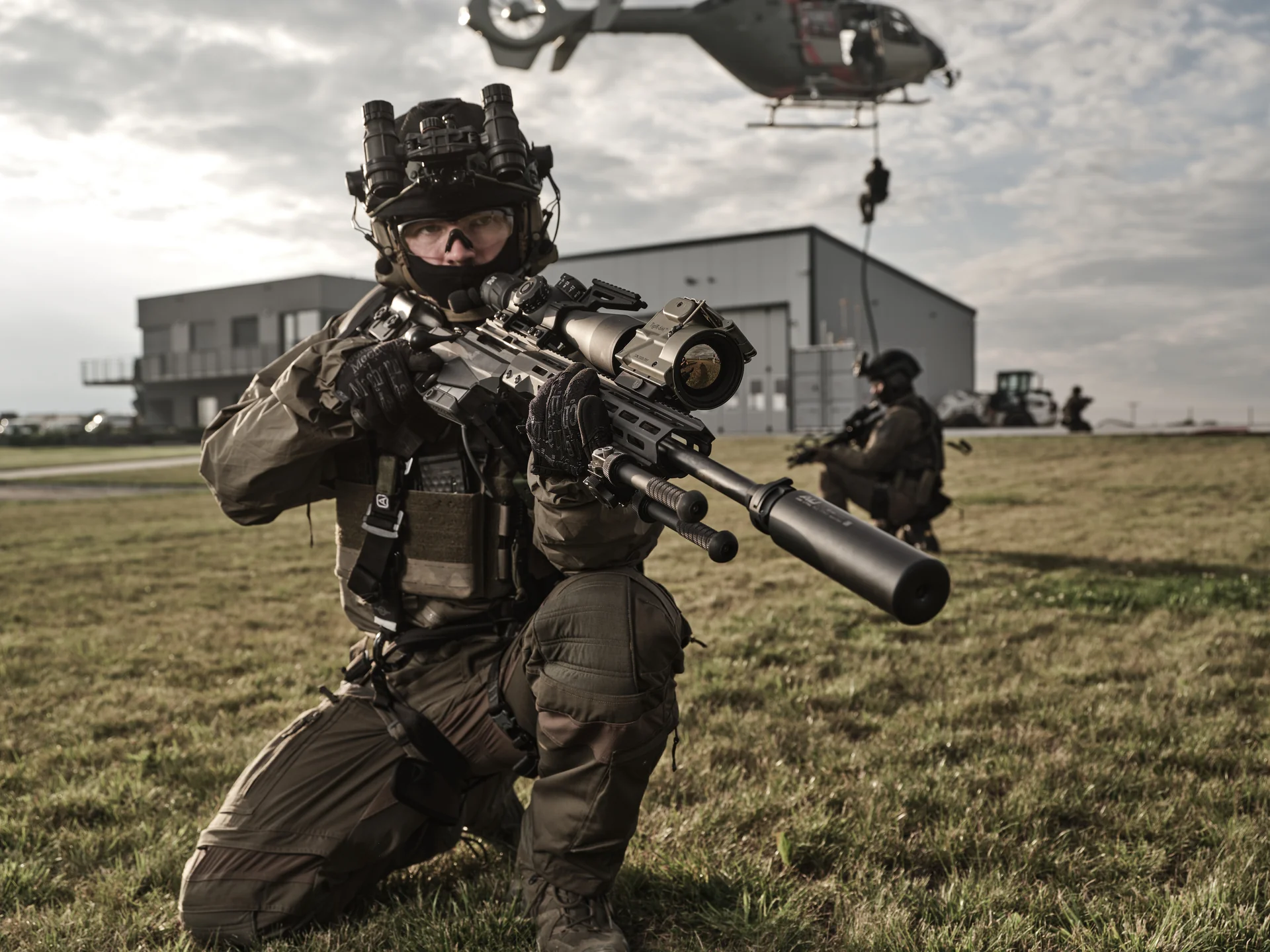 Atlas bipod folded under a long suppressed rifle held by a kneeling operator as a helicopter hovers over an airfield.