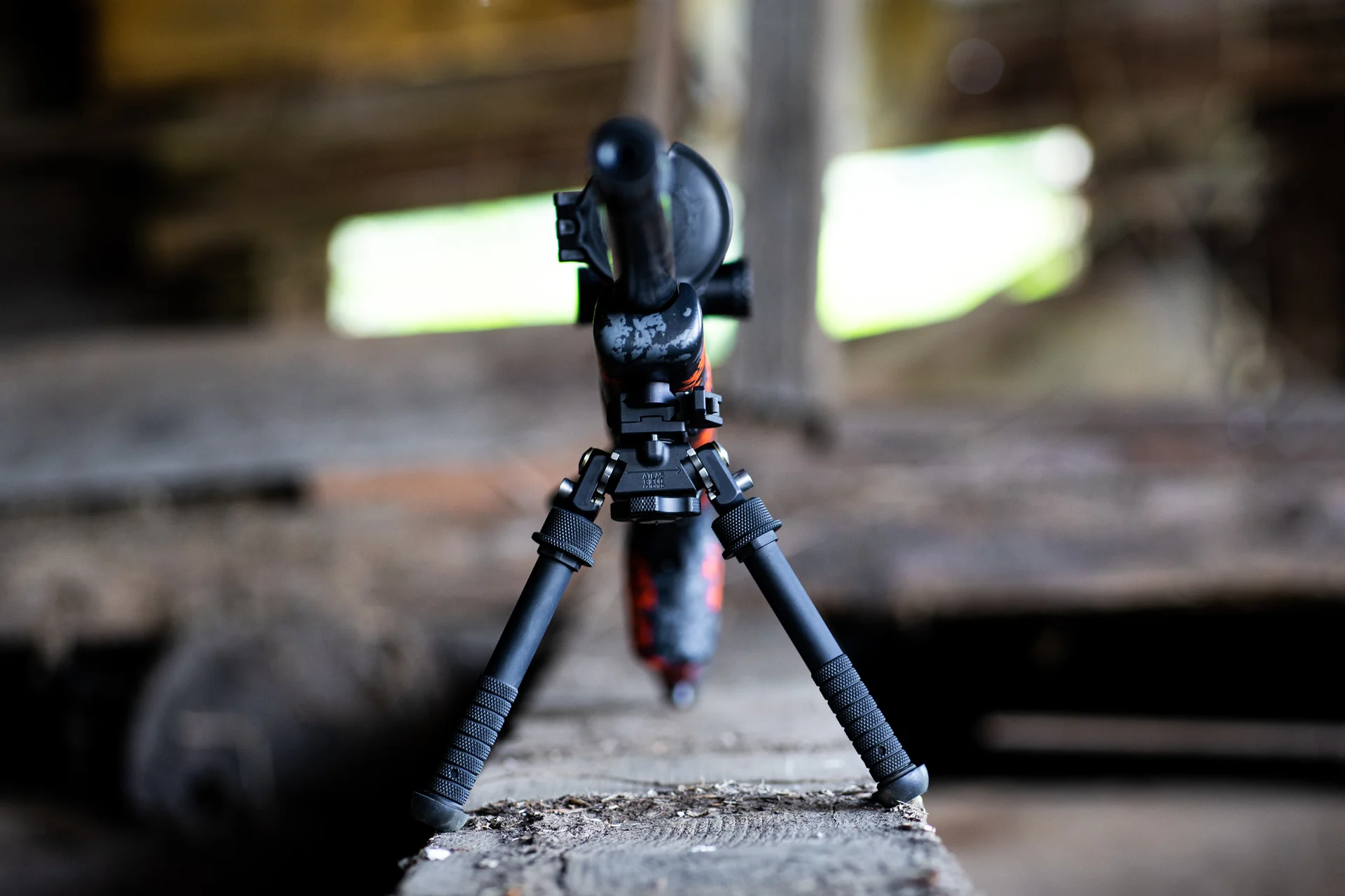 Atlas bipod deployed front-on showing notched aluminum legs and central cant body under a camo rifle in a barn.