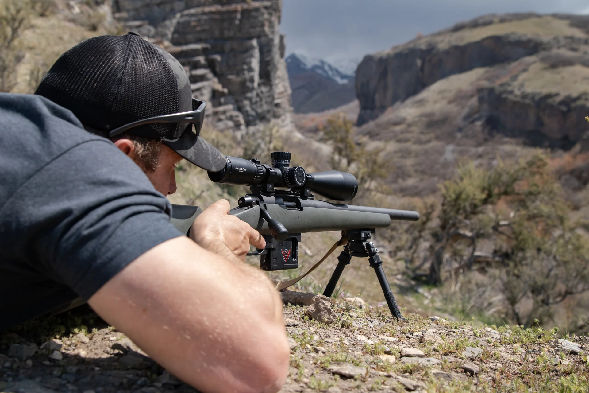 Atlas bipod deployed beneath the rear stock of a green bolt rifle as a kneeling shooter sets up on a canyon overlook.