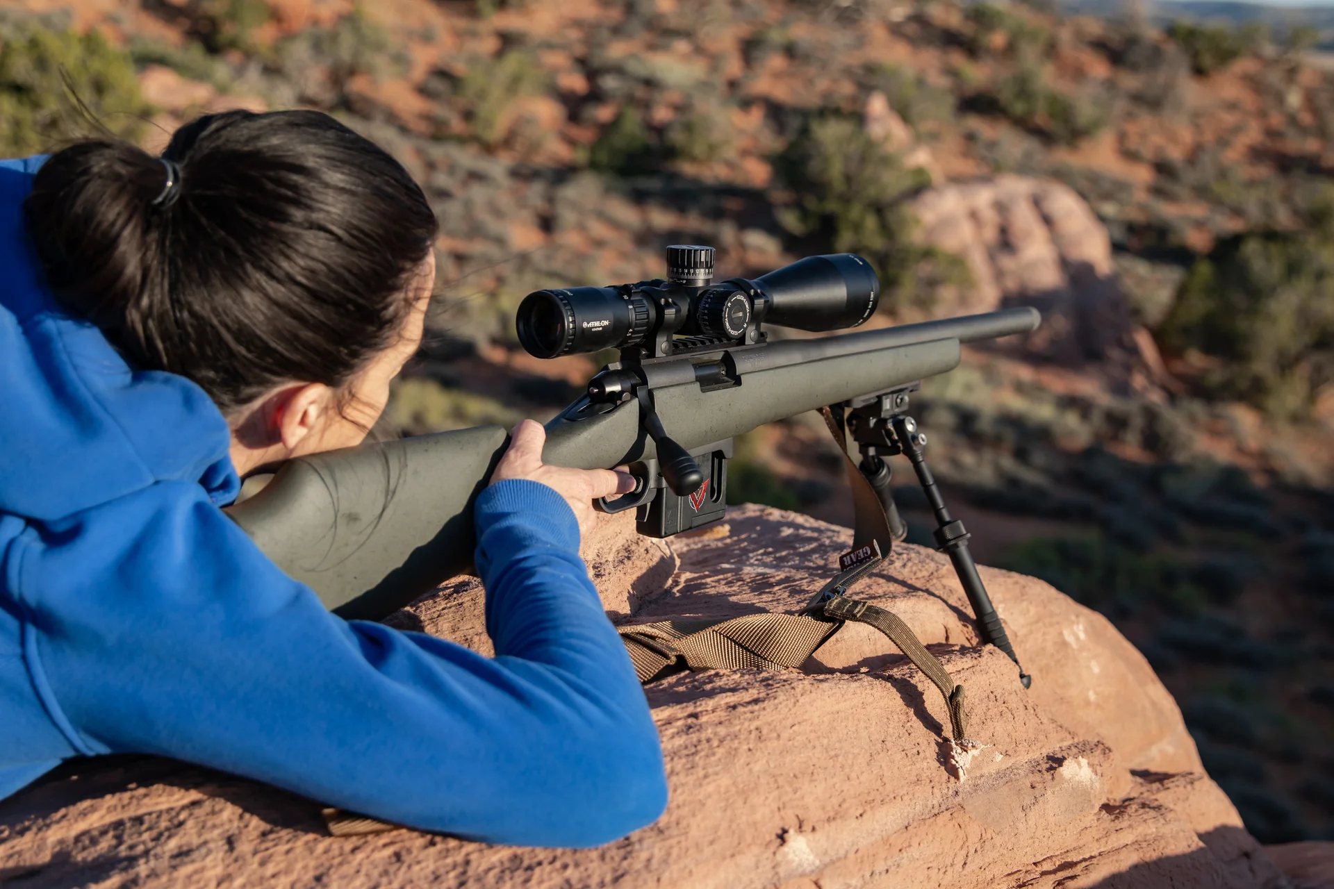 Atlas bipod deployed under the rear stock of a green rifle with a Vortex scope as a prone shooter aims from a mountain ridge.