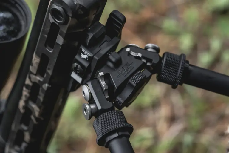 Atlas bipod head detail deployed under a camo rifle showing the pan-and-cant body and notched legs.