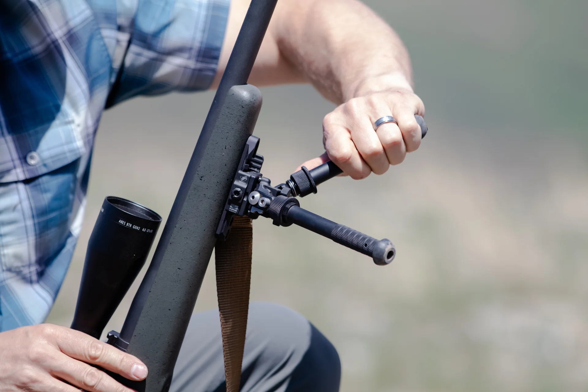 Atlas bipod deployed in closeup as a shooter adjusts a notched leg beneath a rifle with Athlon scope outdoors.