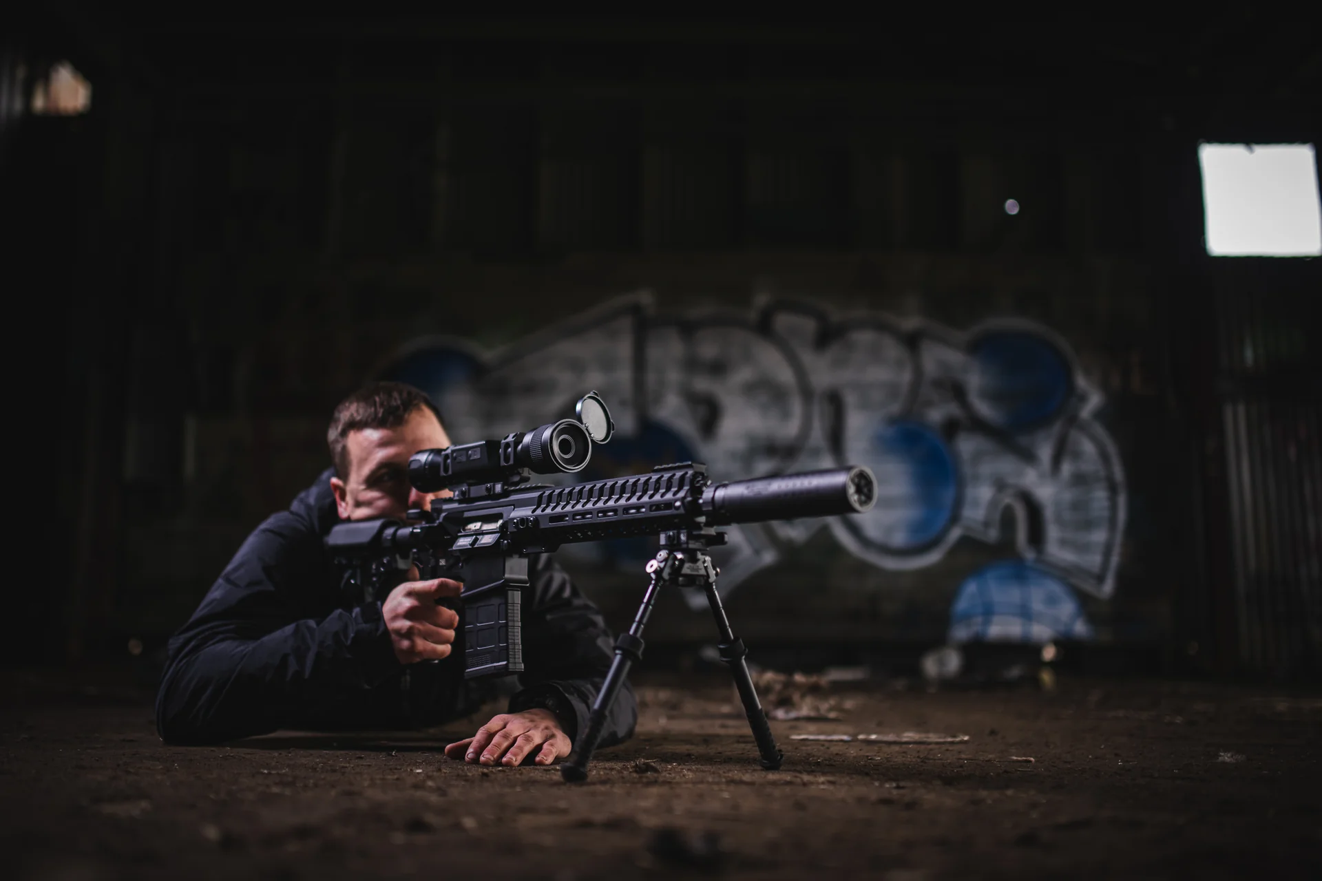 Atlas bipod deployed stabilizing a prone shooter's suppressed AR rifle with thermal optic in a warehouse.