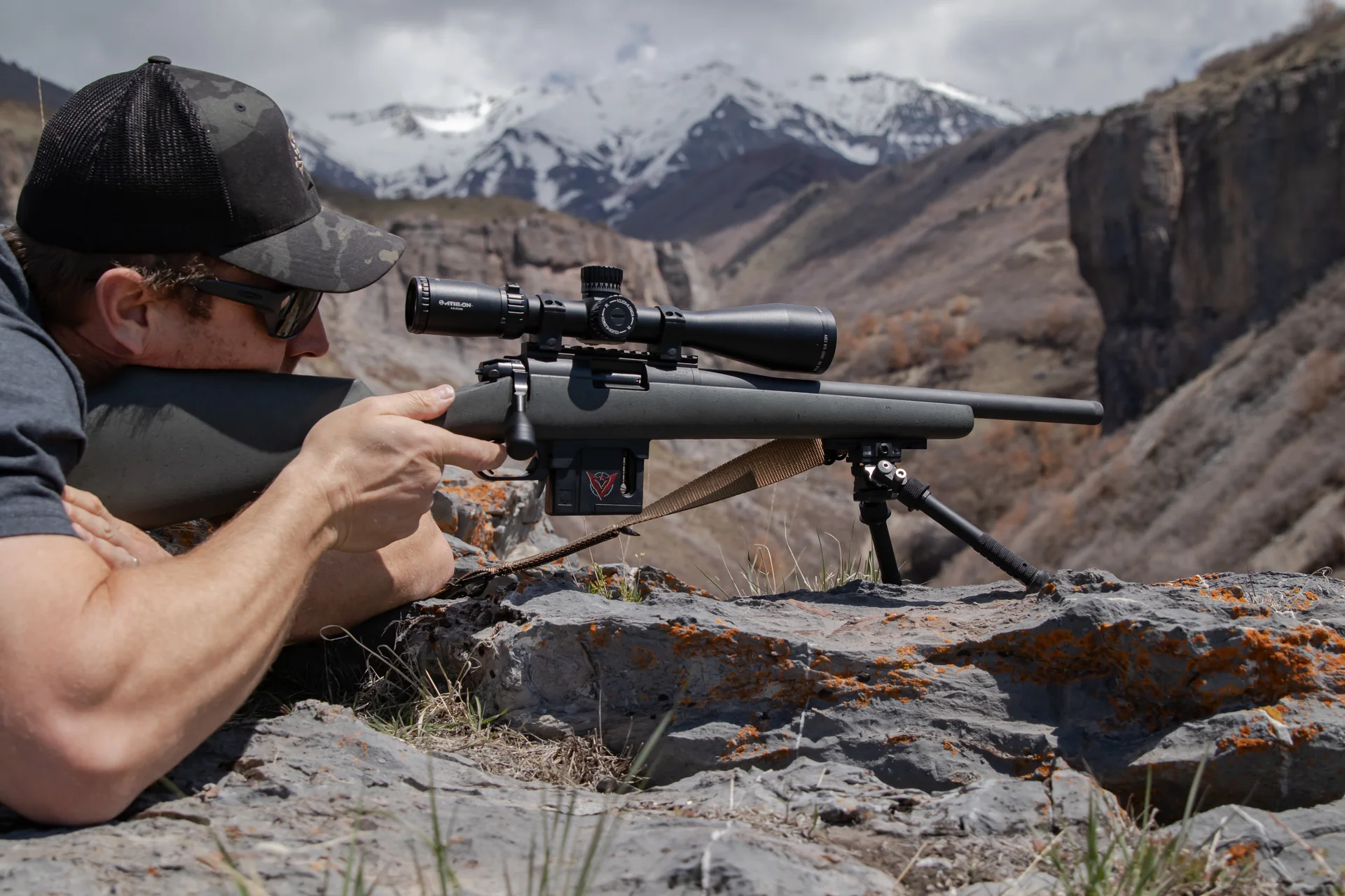 Atlas bipod deployed supports a prone shooter's rifle with Athlon scope against snowy mountain peaks and canyon backdrop.