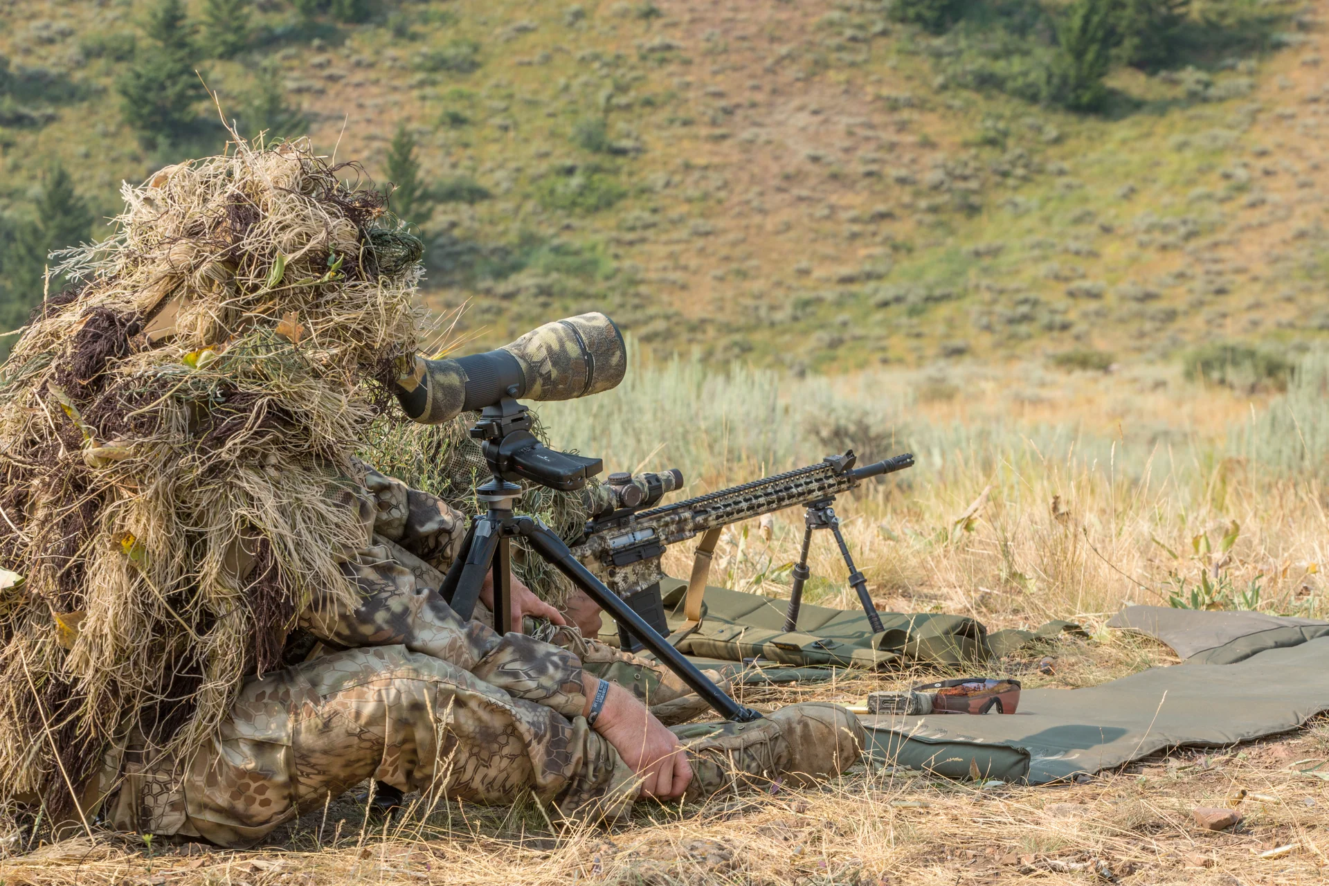 Atlas bipod deployed under a ghillie-clad shooter's rifle as he uses a spotting scope on open sagebrush ground.