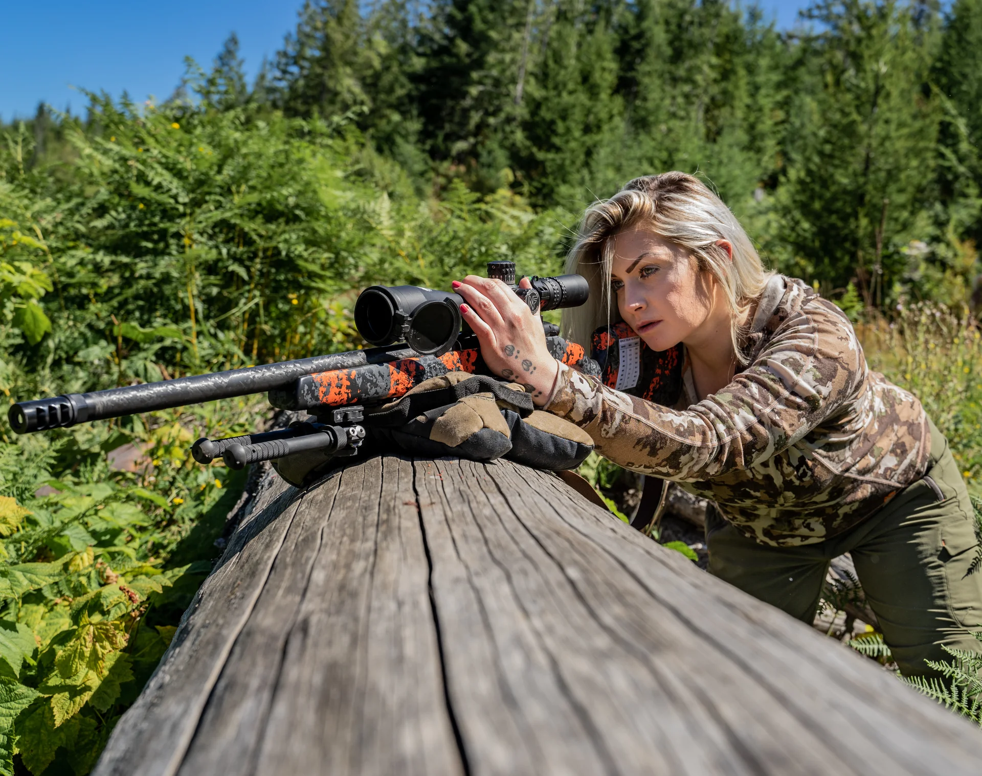 Atlas bipod steadies the rear of a camo bolt-action rifle as a shooter aims over a log in the forest.