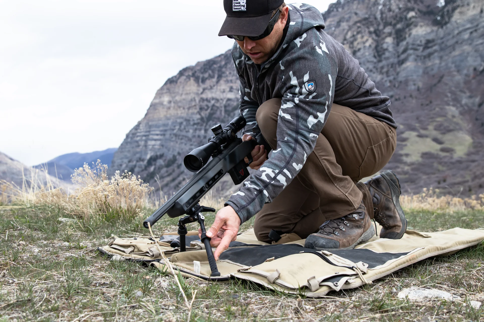 Atlas bipod deployed under the rear stock of a camouflaged bolt rifle as a shooter adjusts it on a mountain ridge.