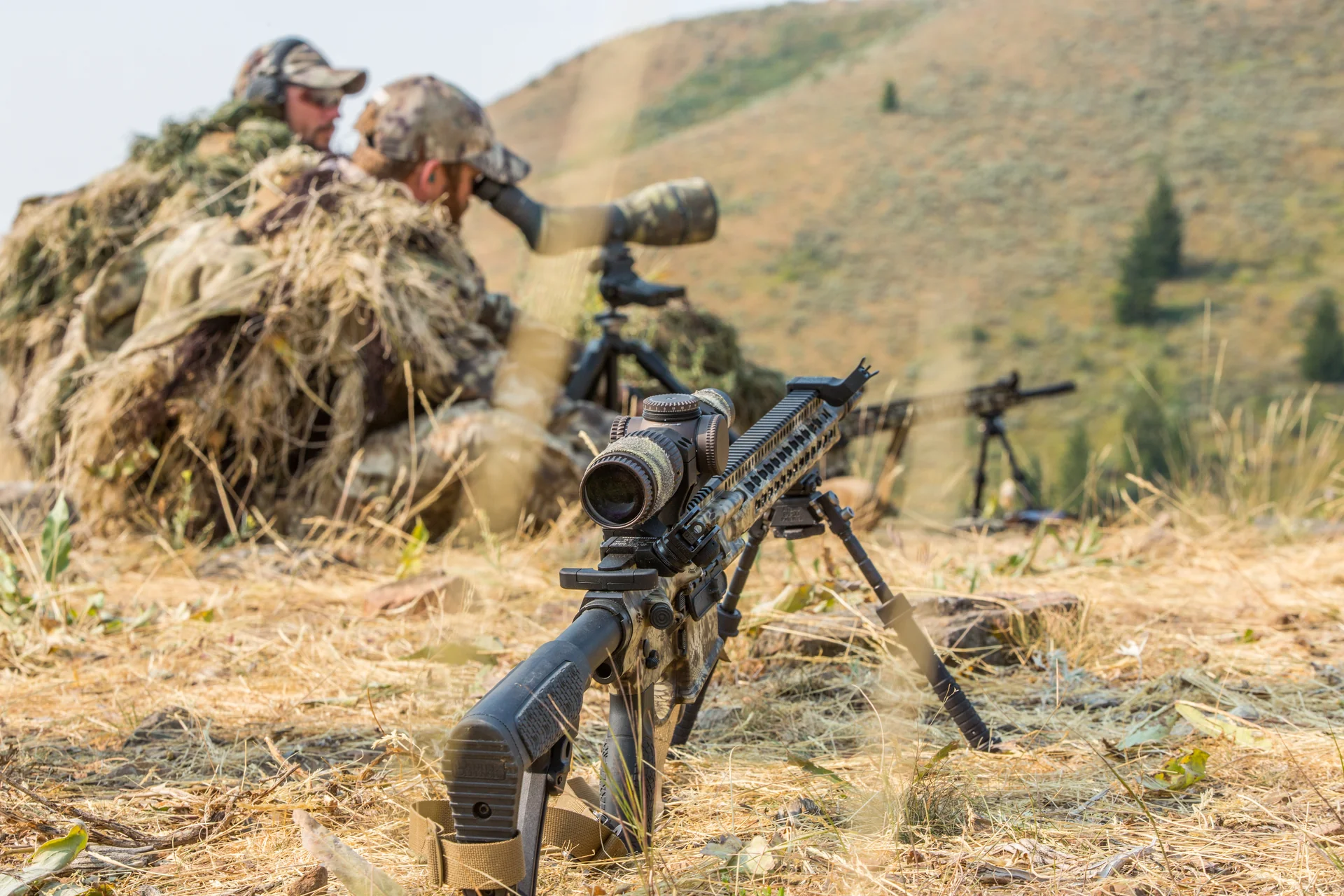 Atlas bipod deployed under a camouflaged AR rifle in sharp foreground focus as a shooter and spotter work the hillside.