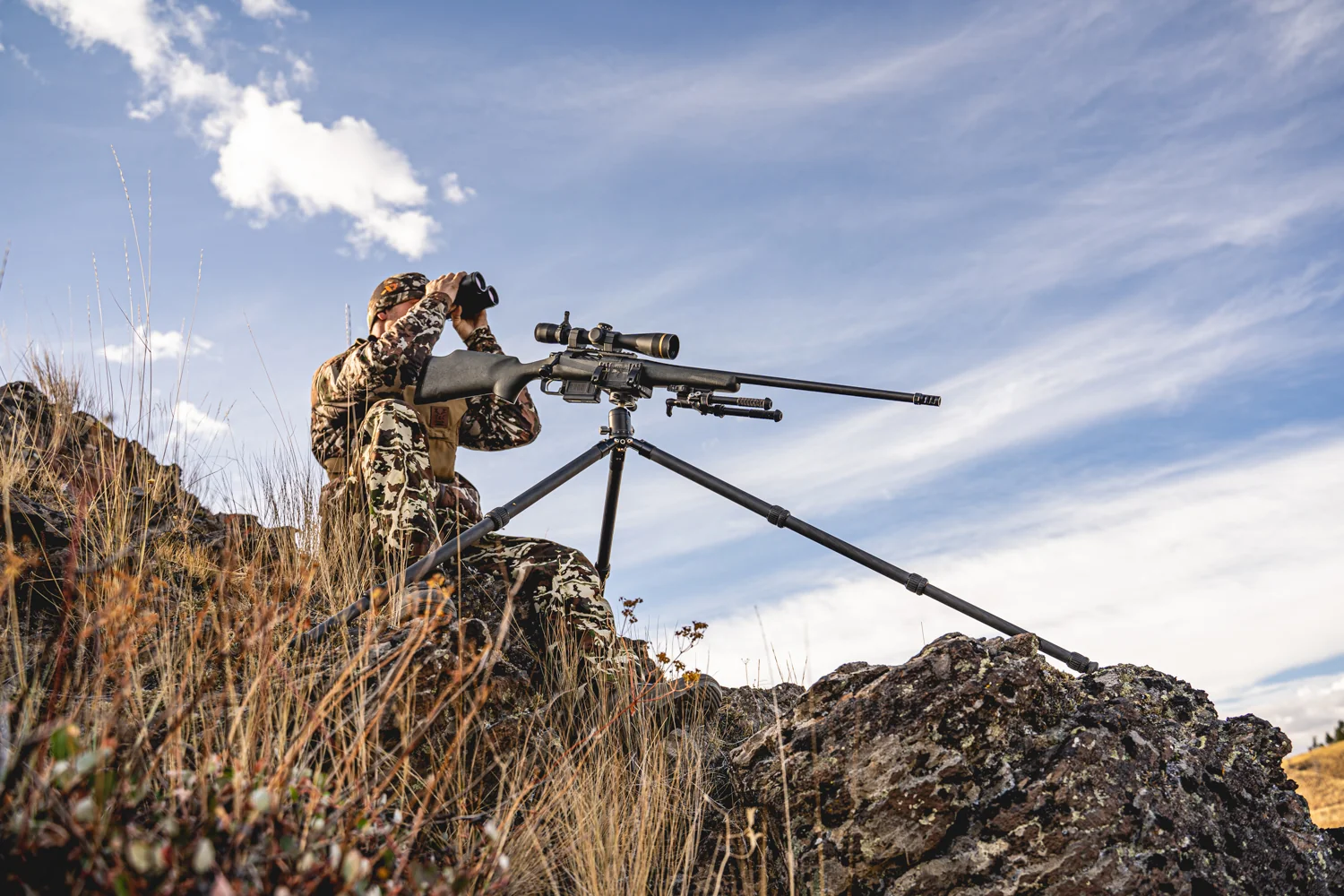 Atlas bipod folded on a tripod-mounted scoped rifle while a hunter glasses across a sunlit ridge.