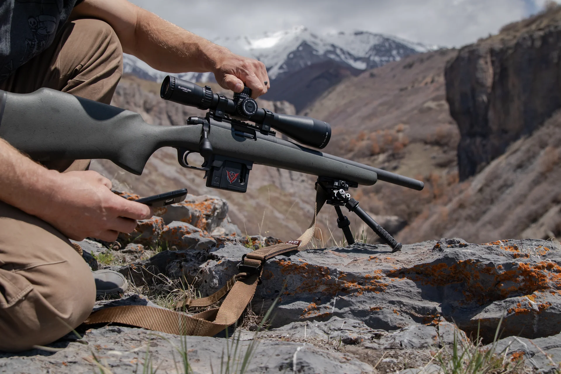 Atlas bipod deployed supports a green-stocked rifle as a shooter adjusts the scope above a rugged mountain canyon.
