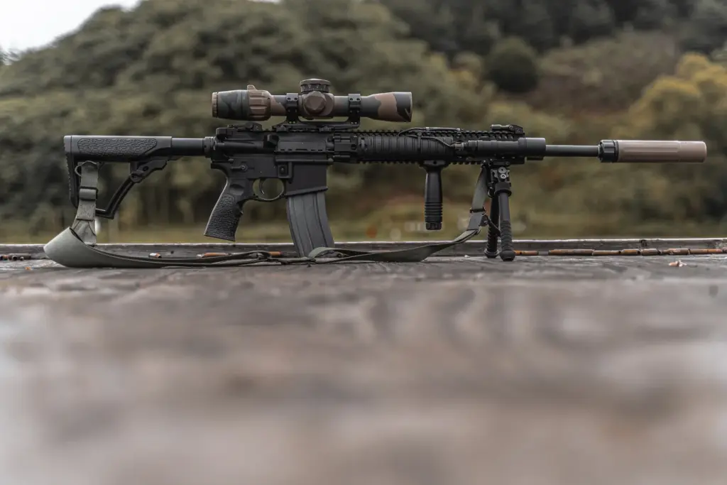 Atlas bipod deployed under a suppressed camo-scoped AR-15 shown in full side profile on a weathered wooden bench.