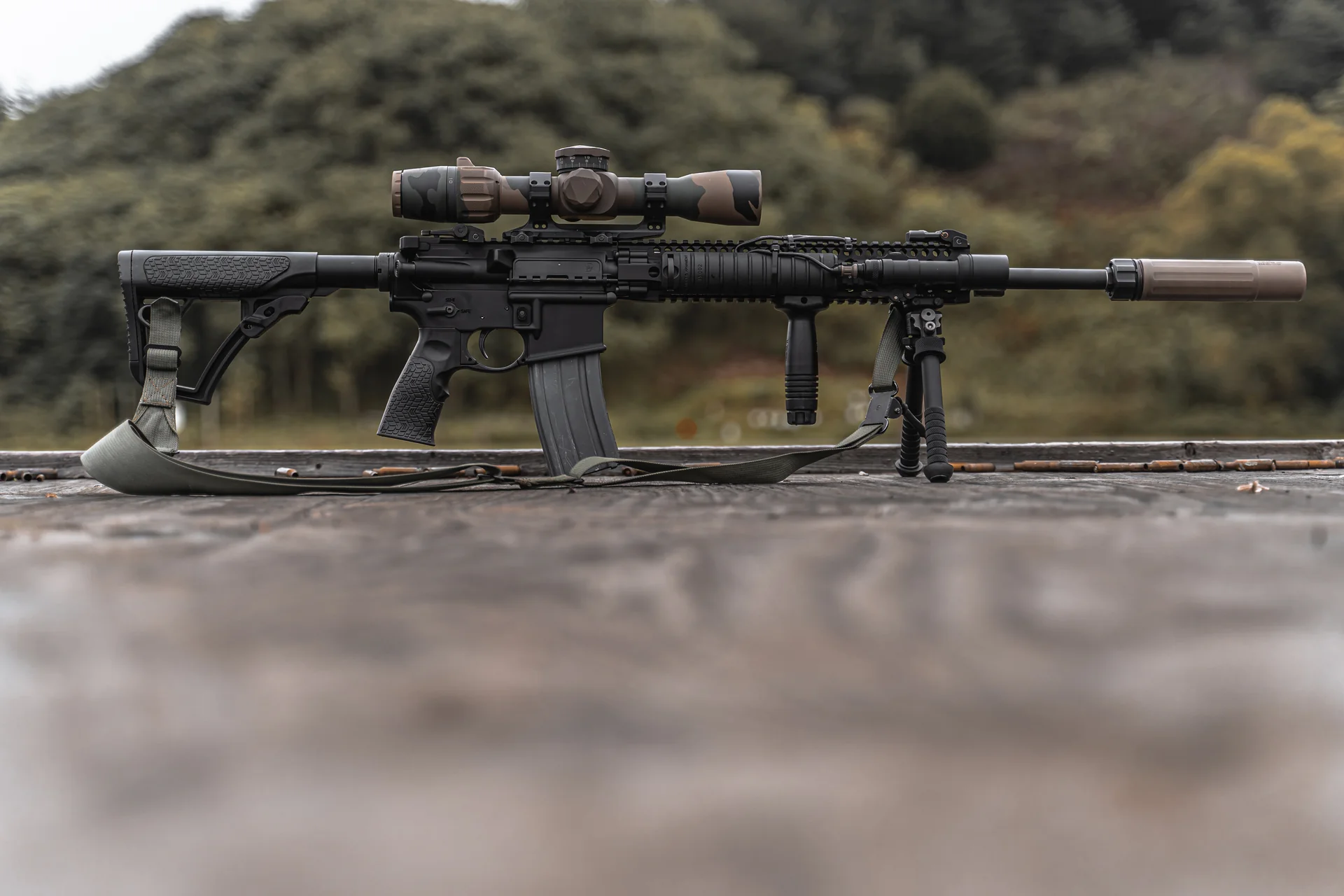 Atlas bipod deployed under a suppressed camo-scoped AR-15 shown in full side profile on a weathered wooden bench.