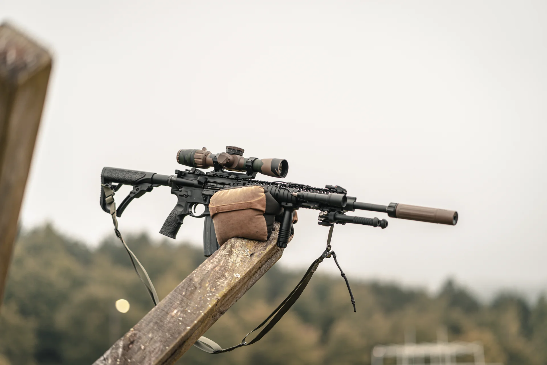 Atlas bipod deployed beneath a suppressed scoped AR-15 resting on a bag atop a fence rail in foggy woods.