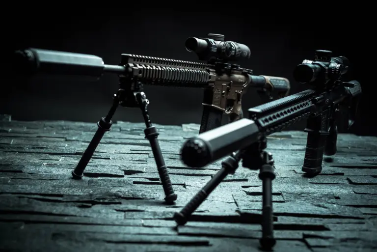 Atlas bipods deployed with notched aluminum legs under a pair of suppressed tactical rifles on slate.