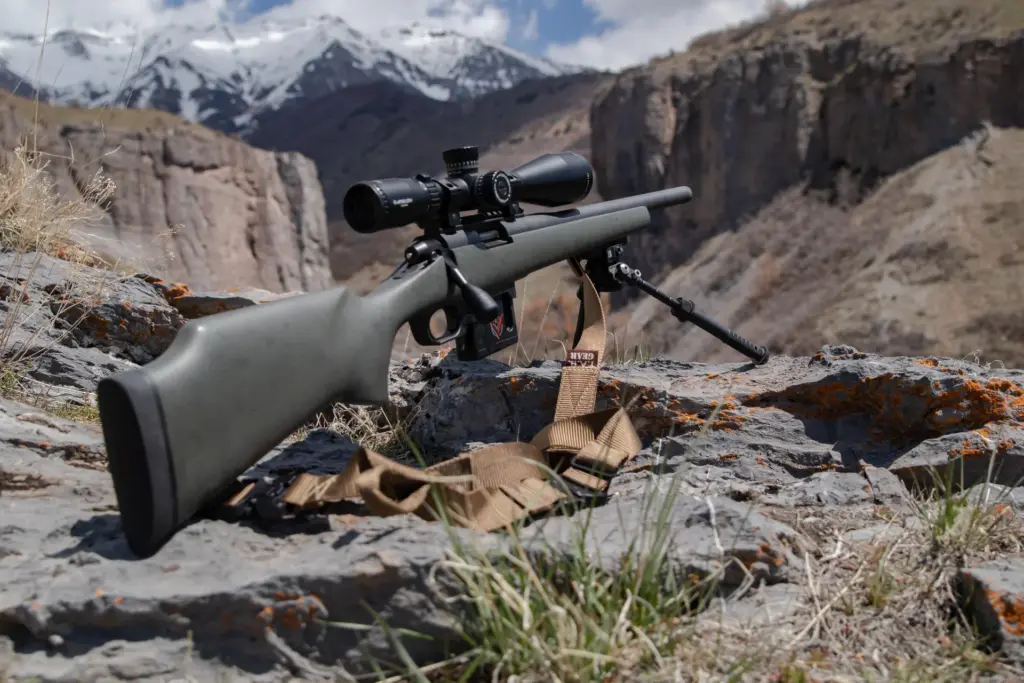 Atlas bipod deployed steadies a scoped green-stocked rifle on lichen-covered rocks framed by a snowy mountain pass.