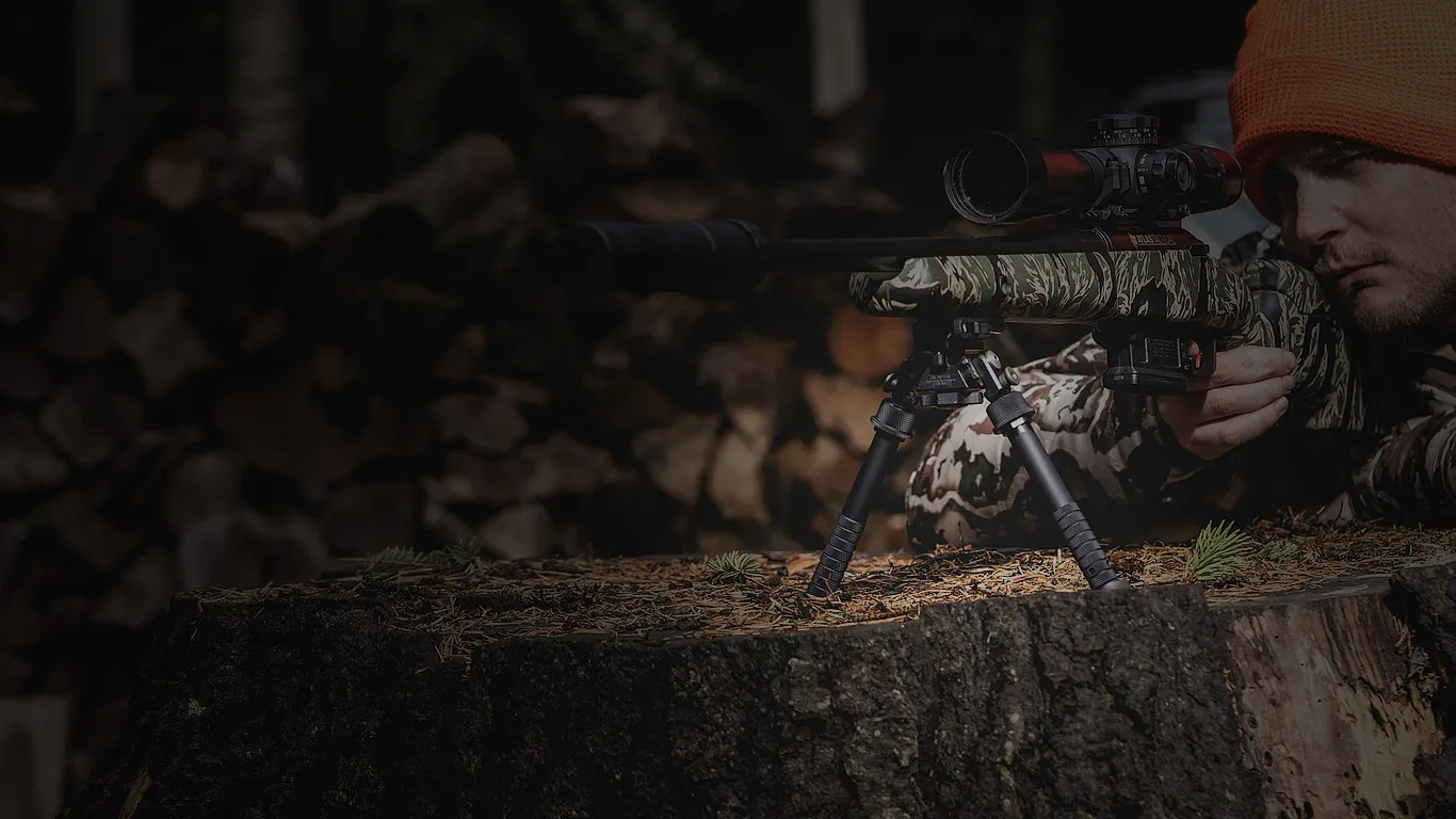 A hunter in camouflage behind a rifle on an Atlas bipod, resting on a tree stump beside a woodpile.